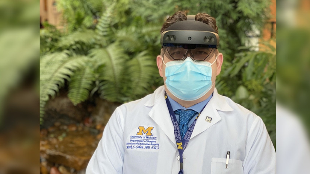 doctor outside in white coat with headset on and blue mask on in front of greenery