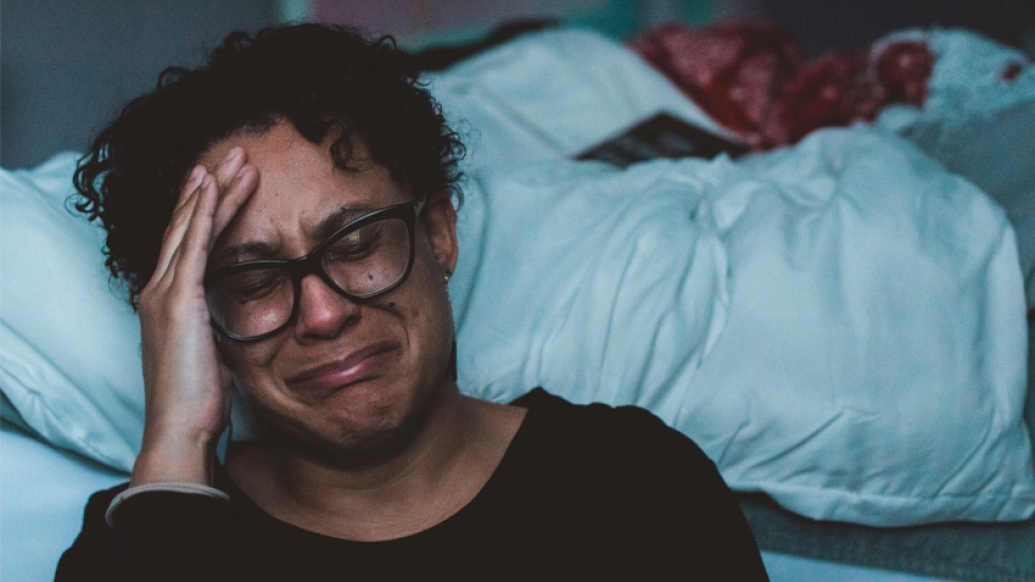 woman crying with glasses next to bed with white blanket 