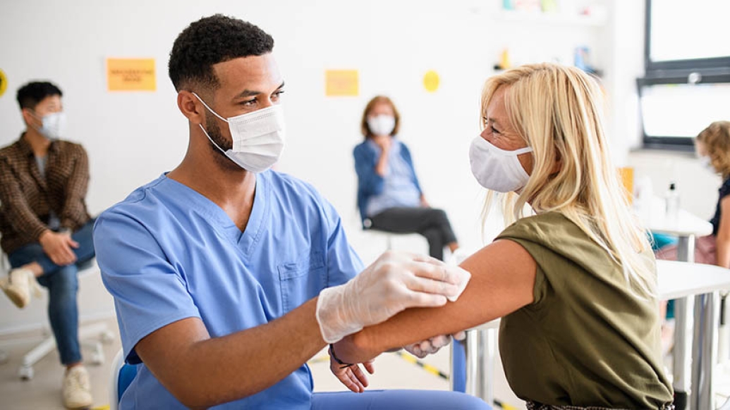 health care worker in blue scrubs and mask giving woman with blonde hair and mask a shot in her arm