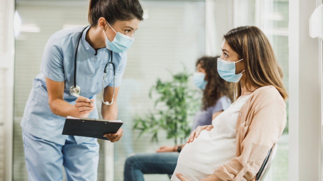 health care worker leaning over to talk to pregnant women in mask sitting